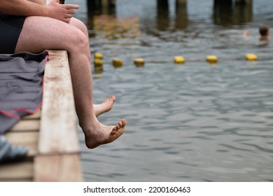 Man Sitting On A Wooden Pier On A Sunny, Summer Day. Only Feet Are Visible. No Shoes. Person Puts His Feet Outside Of Wooden Counstruction And Swings Them Right Above The Water. 