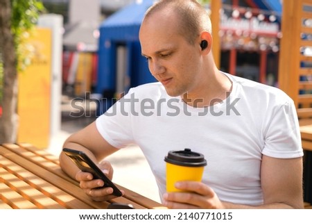 A man is sitting at a table in a cafe drinking coffee from a disposable cup, listening to music through mini in-ear headphones and writing a message in a smartphone smiling sweetly. Lunch break 
