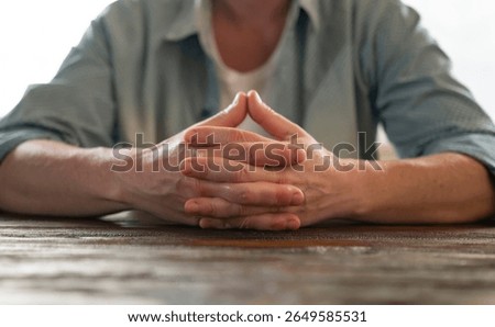 Man sitting at a table with hands clasped, displaying calmness and contemplation in a quiet indoor setting