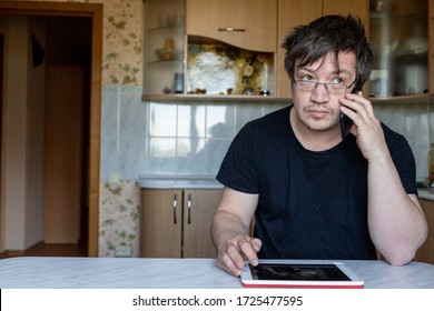 A Man Is Sitting At A Table At Home. Office At Home. The Man Is Talking On The Phone And At The Same Time Looking At The Tablet, Preparing To Record. Not A Model, Just An Ordinary Person. 