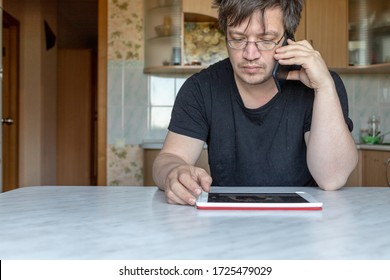 A Man Is Sitting At A Table At Home. Office At Home. The Man Is Talking On The Phone And At The Same Time Looking At The Tablet, Preparing To Record. Not A Model, Just An Ordinary Person. 