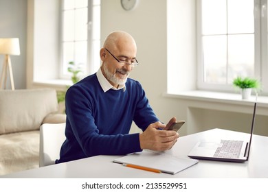 Man Sitting At Table With Mobile Phone. Happy Bald Bearded Senior Man In Glasses Sitting At Desk With Laptop And Notepad, Using Modern Cellphone, Reading News, Looking Up And Studying New Information