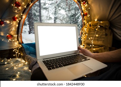 Man sitting in tent decorated with Christmas lights, using laptop. Beautiful top view of Scandinavian landscape, forest covered with snow. Self-isolation and social distancing during holidays