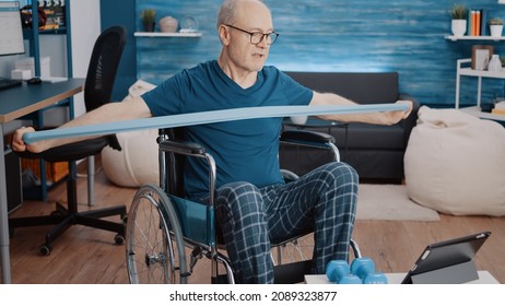 Man sitting in wheelchair and doing exercise with resistance band, looking at video of workout lesson on tablet. Aged person with disability using elastic belt, watching training program