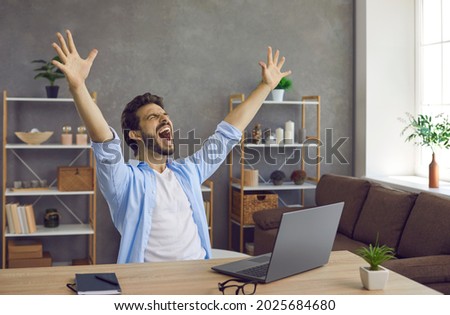 Man sitting at work table with laptop computer feeling excited, raising hands up and screaming. Happy guy finishes business project, learns great news, wins college or university student scholarship