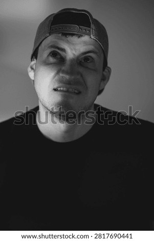 Man with skeptical expression wearing baseball cap. Portrait of confused male looking up with doubtful facial gesture. Cynical emotion or disbelief concept in high contrast black white.