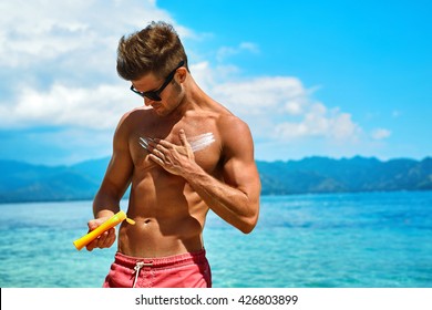 Man Skin Care In Summer. Handsome Male With Sexy Body In Sunglasses Applying UV Protective Sunscreen Lotion Before Sunbathing, Tanning At Beach Using Solar Sun Block Protection Cream For Healthy Tan.