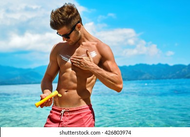 Man Skin Care In Summer. Handsome Male With Sexy Body In Sunglasses Applying UV Protective Sunscreen Lotion Before Sunbathing, Tanning At Beach Using Solar Sun Block Protection Cream For Healthy Tan.