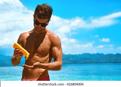 Man Skin Care In Summer. Handsome Male With Sexy Body In Sunglasses Applying UV Protective Sunscreen Lotion Before Sunbathing, Tanning At Beach Using Solar Sun Block Protection Cream For Healthy Tan.