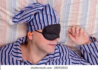 Man Sleeping With A Mask On Eyes. Man In Striped Pajamas Sleeping In Bed With A Night Cap And Eye Protection Blindfold. Above Portrait Dreaming Happy Man With Eye Patch.