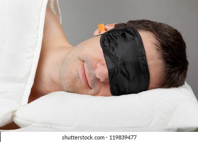 Man With Sleeping Mask Sleep And Earplugs Lying In Bed