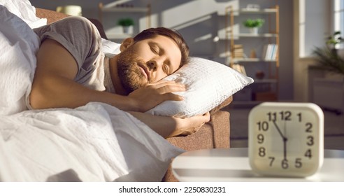Man sleeping peacefully on the sofa in a sunny bedroom or living room with an alarm clock showing 6 AM standing on the table in the foreground on a usual weekday. Morning concept. Banner background - Shutterstock ID 2250830231