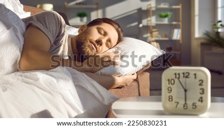 Man sleeping peacefully on the sofa in a sunny bedroom or living room with an alarm clock showing 6 AM standing on the table in the foreground on a usual weekday. Morning concept. Banner background