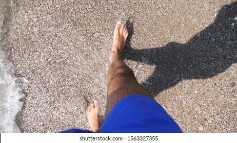 Man Slowly Walking Barefoot On Coast At Sunny Day. Small Waves Splashing Over Feet Of Young Guy Strolling On Sea Pebble Beach. Guy Enjoying Recreation On Seashore. Concept Of Summer Vacation. Slow Mo