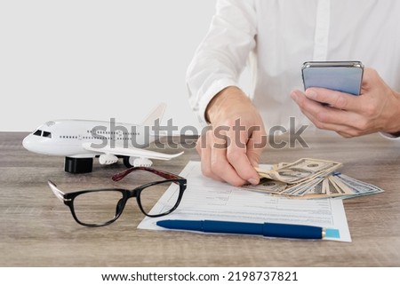 Man with smartphone applying travel insurance form with plane model, glasses and cash money lying on table, studio, light gray background