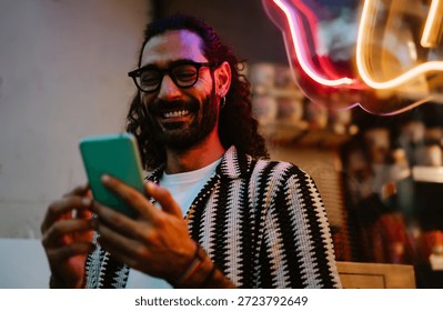 Man smiles warmly while using smartphone near neon-lit window, expressing authentic joy, screen presence and effortless mobile lifestyle connection.