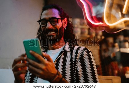 Man smiles warmly while using smartphone near neon-lit window, expressing authentic joy, screen presence and effortless mobile lifestyle connection.