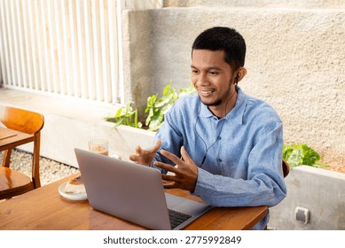 Man smiles while video calling using laptop at outdoor cafe. He wears blue shirt and earphones. Laptop rests on wooden table beside coffee. Green plants and stone wall frame background.
