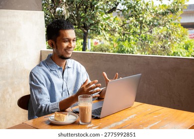 Man smiles while video calling using laptop at outdoor cafe. He wears blue shirt and earphones. Laptop rests on wooden table beside coffee. Green plants and stone wall frame background.