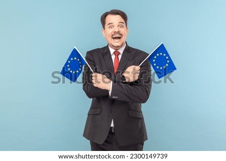 Man smiling broadly and holding flags of European Union, symbol of Europe, EU association and community, wearing black suit with red tie. Indoor studio shot isolated on light blue background.