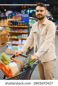 Man smiling at camera while pushing a shopping cart filled with fresh produce and groceries through a supermarket aisle