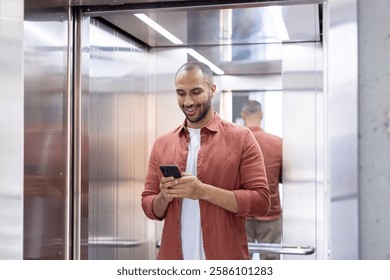 A man is smiling in an elevator while looking at his smartphone, wearing a casual terracotta shirt and a white t-shirt