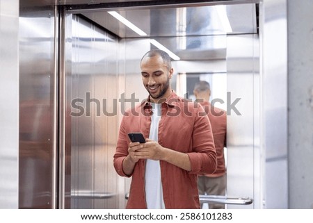 A man is smiling in an elevator while looking at his smartphone, wearing a casual terracotta shirt and a white t-shirt