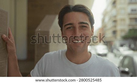 Man smiling with face forward, left hand on doorway, wearing white tshirt and hoop earring on street near building entrance; casual warmth.