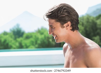 Man smiling and facing left, posing shirtless at pool edge with pool water, copy space. Barechest, male, outdoor, sunlit, turquoise, deck, trees