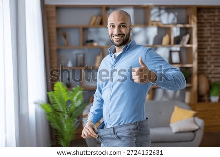 Man smiling and giving thumbs up while pulling out loose jeans to show weight loss success, confident and happy after healthy lifestyle changes at home, feeling fit and motivated