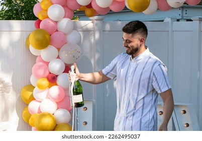 A man smiling and popping a bottle of champaign after she said yes standing under balloons in backyard.