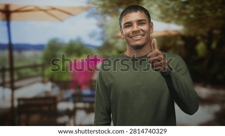 Man smiling and showing thumb while pointing finger toward camera in studio wearing green long sleeve shirt and goatee, casual pose conveys warmth; friendly approval.