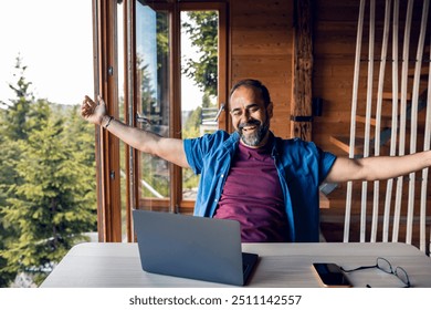 Man smiling and stretching while working on a laptop in a wooden cabin with a view of the forest - Powered by Shutterstock