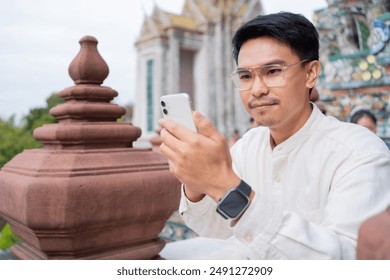 A man is smiling and waving at the camera while holding a white iPhone. He is wearing a watch and glasses