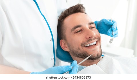 A man is smiling while a dentist works on his teeth. The dentist is wearing blue gloves and holding a pair of dental tools. - Powered by Shutterstock