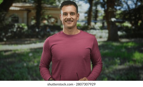 Man smiling while holding glasses in front of a building, wearing a burgundy long sleeve shirt and wristwatch; calm confidence.