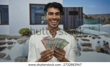 Man smiling while holding rupee banknotes at the sunlit entrance of a modern white building; prosperity.