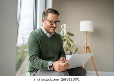 Man smiling while sitting by a window, using a laptop for remote work or a video call in a relaxed, smart-casual home office environment, focused and comfortable