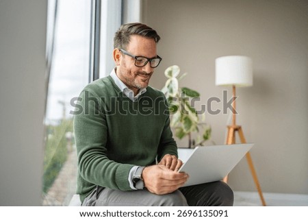 Man smiling while sitting by a window, using a laptop for remote work or a video call in a relaxed, smart-casual home office environment, focused and comfortable