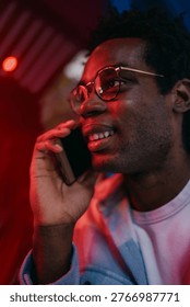 Man smiling while talking on smartphone under intense red lighting, reflecting modern social interaction and the emotional resonance of real-time digital conversations.