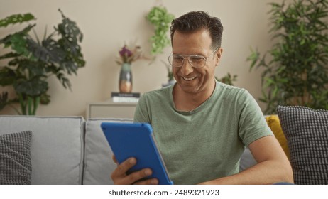 Man smiling while using blue tablet in a cozy living room filled with plants and modern decor - Powered by Shutterstock - Get 15% off with code: PIKWIZARD15