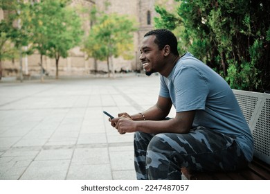 Man smiling while using mobile phone sitting on a bench outdoors.