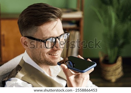 Man smiling while using smartphone for voice recognition in a cozy indoor setting during the day