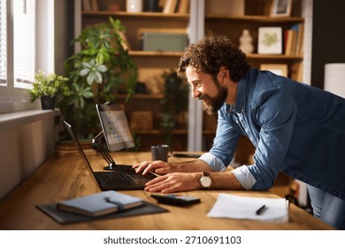 A man is smiling while working on his laptop in a cozy home office. The workspace features natural light, plants, and neatly arranged books on shelves, creating a productive atmosphere.