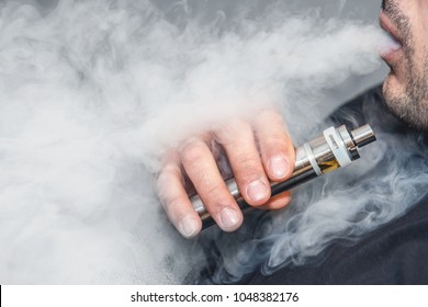 A man smokes an electronic cigarette on a gray background, blowing a stream of smoke. Copy space. - Shutterstock ID 1048382176