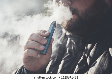 Man smokes new Vape Pod System, inhales and exhales vapor of electronic cigarette, vaping concept, selective focus, toned