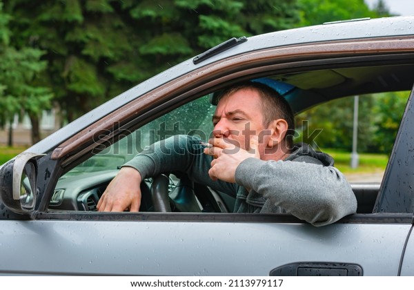A man smokes in the rain while sitting in the car\
through the window