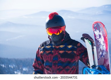 A Man In Snowboarding Gear On The Top Of A Mountain. Young Man Standing With A Snowboard On The Top Of The Mountains