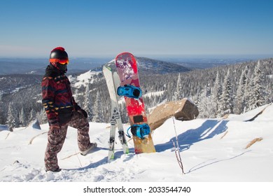 A Man In Snowboarding Gear On The Top Of A Mountain. Young Man Standing With A Snowboard On The Top Of The Mountains