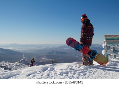 A Man In Snowboarding Gear On The Top Of A Mountain. Young Man Standing With A Snowboard On The Top Of The Mountains.